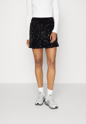 Vrouw draagt zwarte sequin shorts, wit top met lange mouwen, witte sokken en grijze Adidas sneakers, staand tegen een effen achtergrond.