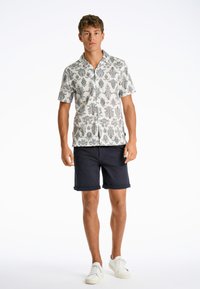 Camisa blanca de manga corta con patrones florales negros, combinada con pantalones cortos azul marino enrollados y zapatillas blancas. Atuendo de verano simple y casual.
