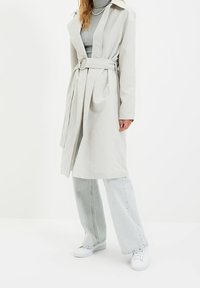 Mujer vistiendo un abrigo trench beige claro con cinturón sobre un suéter de cuello alto gris, combinado con jeans de pierna ancha azul claro y zapatillas blancas.