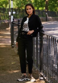 Czarna kurtka zapinana na guziki nałożona na koszulkę z grafikiem, w zestawieniu z dopasowanymi czarnymi spodniami i beżowymi sneakersami z ciemnymi akcentami oraz masywną podeszwą.