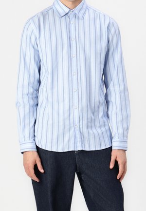 Man wearing a light blue long-sleeve shirt with vertical stripes and dark blue pants, standing against a plain white background.