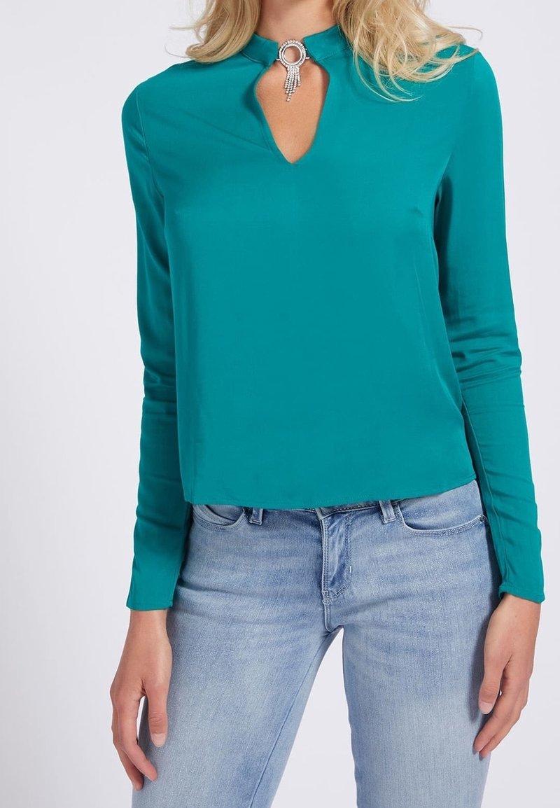 Blusa lunga teal con scollatura a chiave e dettagli in argento. Indossata con jeans fitted azzurri. Tessuto morbido, vestibilità su misura.