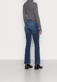 Mörkblå utsvängda jeans med medelhög midja, bakfickor och en detalj i brunt läder. Modellen bär en grå, ribbad polotröja.