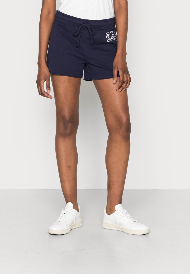 Personne portant un short bleu marine avec cordon et logo "GAP" sur le côté gauche, des baskets blanches et un haut blanc, debout sur un sol gris.