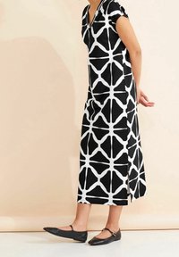 Svart och vit mönstrad klänning med korta ärmar, V-ringning och medellång längd, kombinerad med svarta platta skor mot en neutral bakgrund.