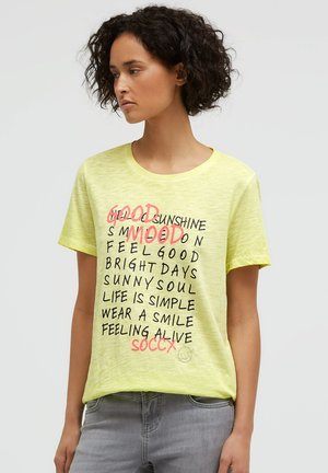 Woman with short curly hair wearing a light yellow T-shirt with motivational text in black and coral lettering, paired with grey jeans.