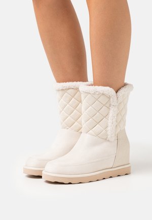Stivali invernali color crema con fodera trapuntata e risvolti in peluche indossati su gambe nude contro uno sfondo bianco.