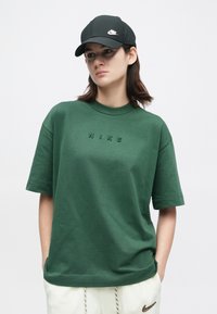 Νέος άνθρωπος φορώντας ένα σκούρο πράσινο μπλουζάκι Nike, άσπρα παντελόνια Nike και μαύρο καπέλο Nike, ενάντια σε ένα απλό φωτεινό φόντο.