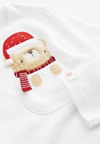Gestickter Bär, der einen roten Weihnachtsmannhut und einen gestreiften Schal trägt und aus einer kleinen weißen Tasche auf einem weißen Stoff hervorblitzt.