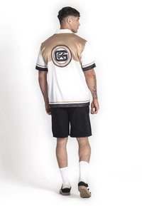 Polo de manga corta en blanco y dorado, con detalles en negro y un logo circular en la parte posterior. Combinado con pantalones cortos negros y calcetas blancas.