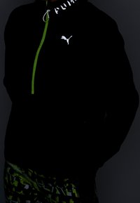 Henkilö, jolla on päällään musta Puma-takki, jossa on vihreä vetoketju, ja vihreät, mustat ja valkoiset kuvioidut urheiluleggingsit tummalla taustalla.