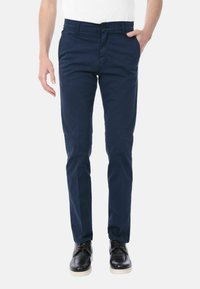 Pantaloni di cotone blu navy con vestibilità slim, dotati di chiusura frontale con bottone, tasche laterali e una texture liscia. Accompagnati da scarpe nere.