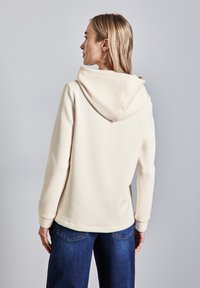 Felpa crema con una texture morbida, dotata di un ampio cappuccio, maniche lunghe e un orlo leggermente curvo, abbinata a jeans blu scuro.