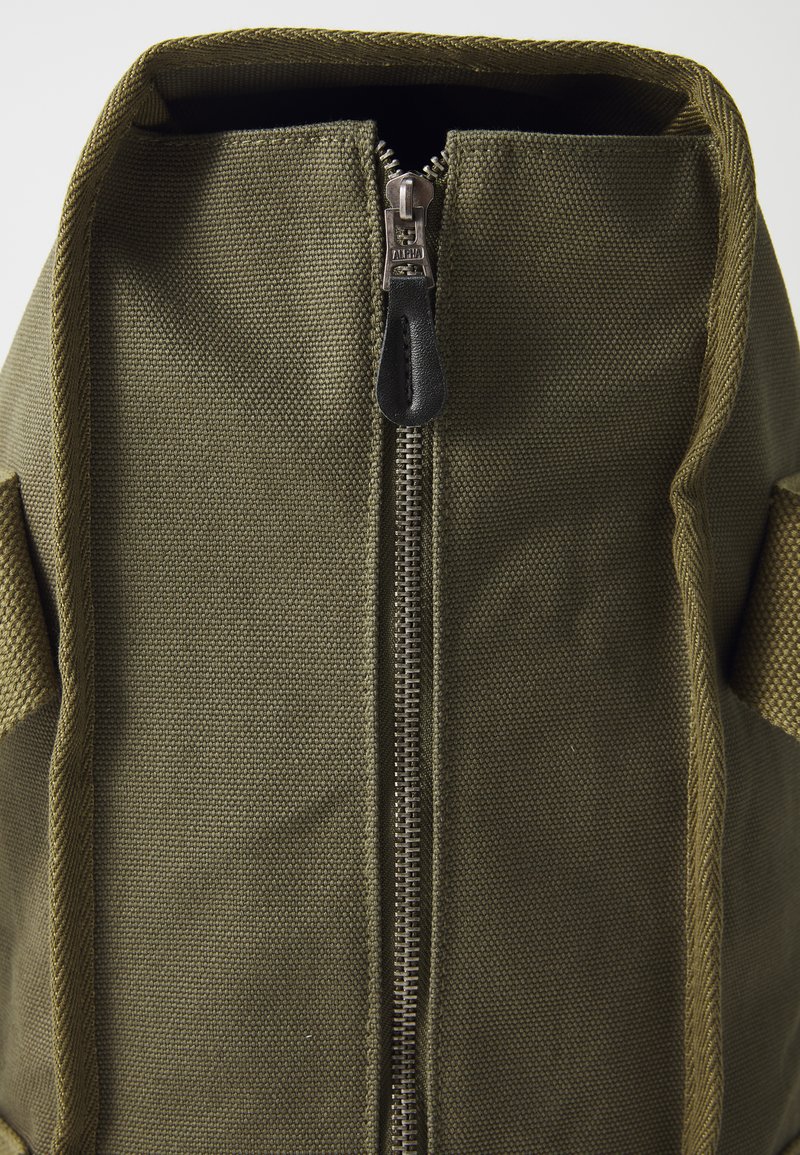 Zielonooliwkowy plecak z kanwy z centralnym zamkiem błyskawicznym, tekstura tkaniny i bocznymi kieszeniami. Trwały materiał z prostym, funkcjonalnym designem.