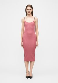 Γυναίκα που φοράει αμάνικο, εφαρμοστό, κοντό φόρεμα σε σκούρο ροζ με ριμπ υφή, με λουριά αλυσίδας, και μαύρες μυτερές γόβες, στέκεται μπροστά σε λευκό φόντο.