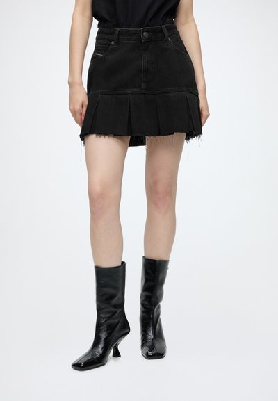 Femme portant une mini-jupe plissée noire à ourlet effiloché et des bottes noires brillantes à talons mi-mollet, sur un fond uni.