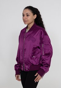 Paarse satijnen bomberjack met ritssluitingen aan de zakken, geribde manchetten en geborduurde details. Soepele textuur, ontspannen pasvorm.