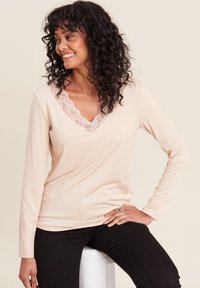 Langärmliges, hellbeiges Oberteil mit V-Ausschnitt und Spitzenbesatz am Ausschnitt, kombiniert mit enganliegenden schwarzen Hosen. Weicher Stoffeindruck.