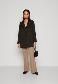 Trench coat nero doppiopetto con grandi bottoni, pantaloni beige svasati e una piccola borsa nera con un accento floreale. Tessuto dalla consistenza liscia.