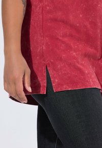 Rode, licht gestructureerde top met zijsplitten, gecombineerd met zwart, strak zittende jeans. De stof heeft een subtiele glans en variatie.