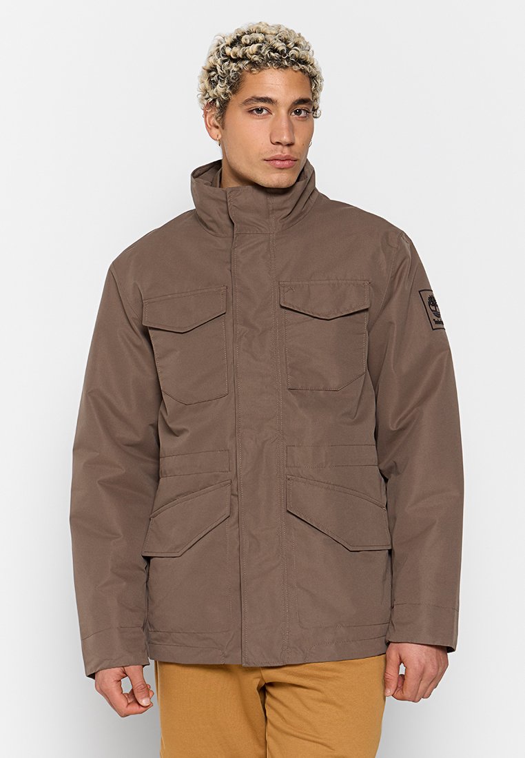 Giacca impermeabile marrone con colletto alto, due tasche sul petto, tasche laterali e un'etichetta nera con logo sul braccio sinistro. Presenta una texture solida.