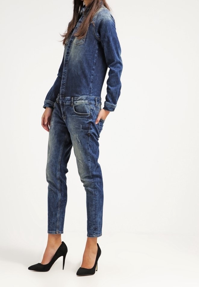 Femme portant une veste en jean bleue délavée et un jean assorti avec des chaussures à talons noirs, debout devant un fond uni.