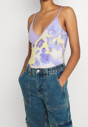 Personne portant un haut sans manches violet et jaune à motif avec de fines bretelles et un pantalon en jean bleu avec de grandes poches devant.