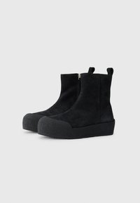 Bottines en suédine noire avec une semelle en caoutchouc, dotées d'une fermeture éclaire sur le côté et de languettes pour un enfilage facile. Texture lisse avec un design à bout rond.