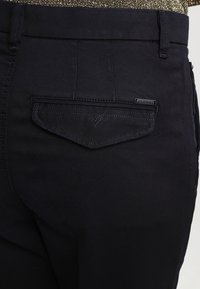 Svarta denimjeans med en bakficka som har ett V-format sömmönster och en liten "Diesel"-etikett. Tyget är slätt och matt.