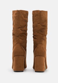 Stivali in suede marrone alti fino al ginocchio con tacco a blocco, texture arricciata, finitura liscia e punta arrotondata, progettati per una vestibilità aderente attorno al polpaccio.