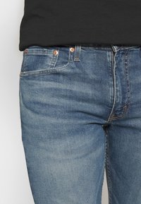 Blaue Jeans aus Denim mit klassischem Schnitt, versehen mit Kupferknöpfen, Vordertaschen und einer verwaschenen Textur.