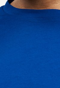 Κοντινή λήψη του λαιμού και του άνω τμήματος του στήθους ενός ατόμου που φοράει μονόχρωμο μπλε μπλουζάκι με στρογγυλή λαιμόκοψη, με ορατή υφή υφάσματος.