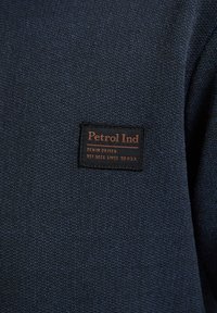 Tessuto testurizzato blu navy con un'etichetta intrecciata nera e marrone che mostra la scritta "Petrol Ind" e un testo aggiuntivo sul denim dal 1989.