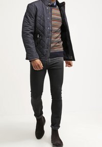 Veste matelassée bleu marine avec fermeture éclair, pull à motif en dessous, associée à un jean noir slim et des bottines marron foncé.