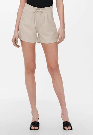 Shorts beige avec un cordon de serrage à la taille, des poches latérales et des coutures décoratives le long des côtés, associés à des sandales noires à enfiler.