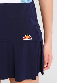 Falda deportiva azul marino con pliegues, que presenta un parche de logo blanco y naranja en la cintura. Fabricada con una tela flexible y suave.