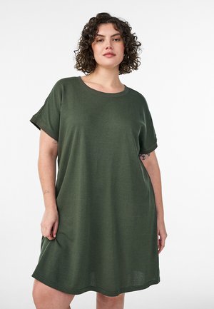 Woman with curly hair wearing loose olive green short-sleeve dress, standing against a plain white background.