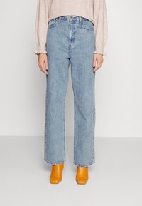 Femme portant un jean droit bleu clair, un pull beige et des bottines orange à bout carré, debout sur un sol gris.