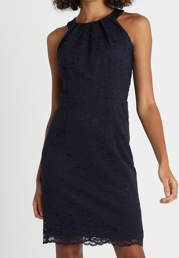 Robe noire en dentelle sans manches avec encolure halter, taille ajustée et ourlet festonné portée par une personne debout sur un fond uni.