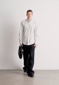 Λευκό πουκάμισο με κουμπιά, σκούρο τζιν με φαρδιά μπότα και μαύρη τσάντα χειρός. Μοντέλο που περπατά με ίσια στάση σώματος σε ουδέτερο φόντο.