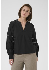 Blouse noire avec un décolleté en V, des manches bouffantes avec des bordures blanches et des détails de plis. Associée à un pantalon texturé beige clair.