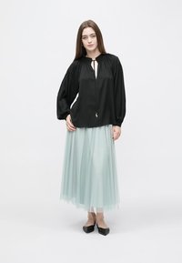 Blusa negra con escote en V profundo y cordón ajustable, combinada con una falda larga de tul en un tono menta claro. La modelo lleva zapatos planos negros.