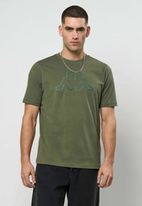 Mann med kort mørkt hår, iført olivengrønn t-skjorte med blått logo og sølvkjede, stående mot en ensfarget lys bakgrunn.