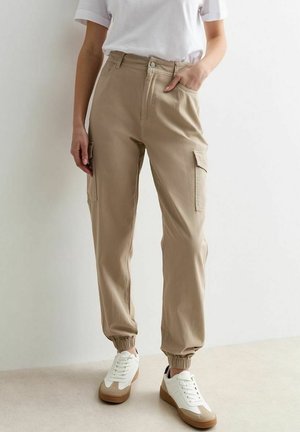 Personne portant un pantalon jogger cargo beige, des baskets blanches avec des semelles marron, et une chemise blanche à manches courtes, debout contre un mur uni.