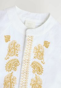 Chemise blanche pour bébé avec broderie paisley dorée, dotée d'un col rond et d'une fermeture à boutons. Texture douce avec un motif quadrillé subtil.