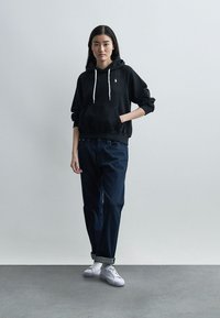 Νεαρή γυναίκα στέκεται με το δεξί χέρι στην τσέπη, φοράει μαύρη κουκούλα, σκουρόχρωμα τζιν με γυρισμένα μπατζάκια και λευκά αθλητικά παπούτσια, πάνω σε απλό ανοιχτό φόντο.