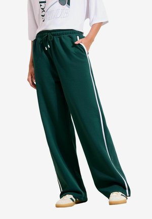 Osoba nosiaca tmavozelené nohavice s širokými nohavicami a bielymi pruhmi na bokoch, biele tričko s grafikou a biele tenisky s čiernymi pruhmi.