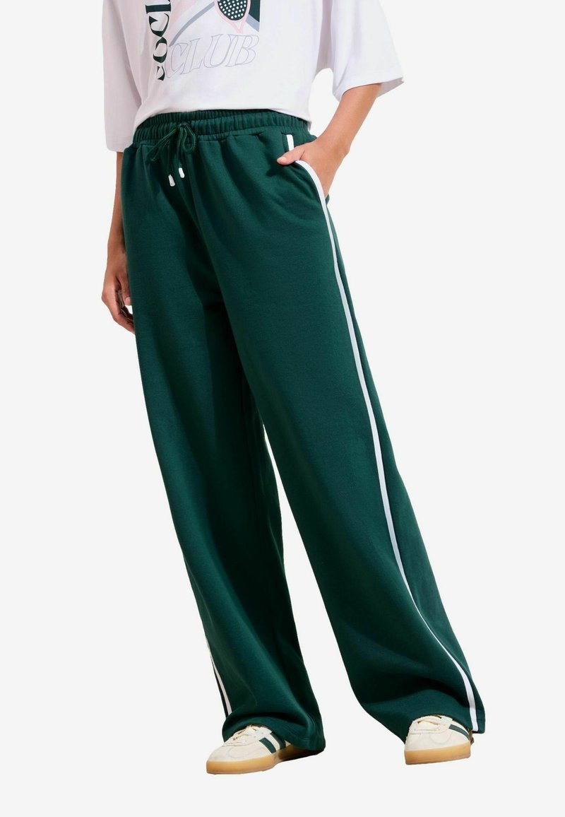 Osoba nosiaca tmavozelené nohavice s širokými nohavicami a bielymi pruhmi na bokoch, biele tričko s grafikou a biele tenisky s čiernymi pruhmi.