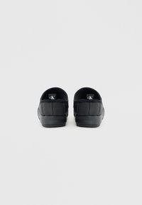 Zapatillas negras sin cordones con forma redondeada, textura acolchada en la parte superior y suela de goma plana. Etiqueta de la marca visible en el talón interior.