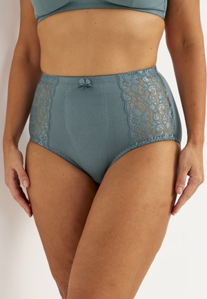 Frau trägt hoch taillierte türkisfarbene Unterwäsche mit Spitzeneinsätzen an den Seiten und einer kleinen Schleife vorne, gezeigt vom mittleren Oberkörper bis zum oberen Oberschenkel.